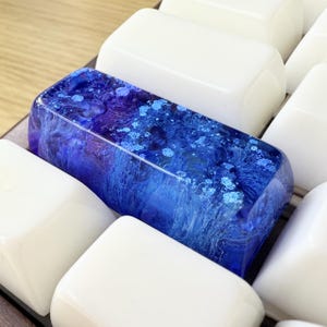 May include: A rectangular mechanical keyboard keycap featuring a deep blue and purple resin design with swirling patterns and embedded glitter particles, positioned among several plain white, smooth-surfaced keyboard keycaps.