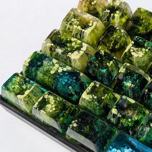 May include: A tray of artisan keycaps in various shades of green and blue. The keycaps have a translucent, resin-like appearance with embedded designs. Some keycaps have a speckled pattern, while others have a marbled effect. The keycaps are arranged in a grid.