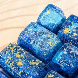 May include: A set of translucent blue resin mechanical keyboard keycaps featuring a swirling pattern of deep navy, bright azure, and metallic gold paint splatters that resemble a starry night sky with textured, glossy surfaces.