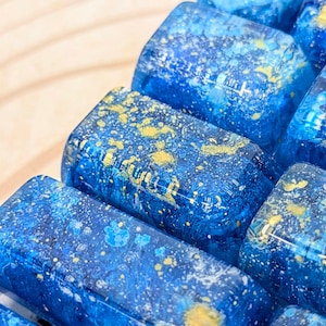 May include: Close-up of several rectangular, blue resin keycaps with gold flecks. The keycaps have a layered, galaxy-like design. The background is a light wood surface.