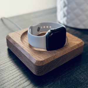 May include: A white Apple Watch with a white sport band is on a wooden charging stand. The stand is on a dark wood surface.
