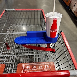 以下が含まれることがあります： 赤いストラップが付いた青いショッピングカートのドリンクホルダー。ストロー付きのコカ・コーラカップが入っています。カップは白地に赤いコカ・コーラのロゴ入り。カートは赤とシルバーです。