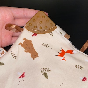 Könnte beinhalten: Ein beiger Baby-Beißring mit einem braunen Silikon-Beißring. Der Beißring hat einen Bär- und Fuchs-Druck mit braunen Pilzen und grünen Blättern.