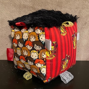 May include: A red and black fabric cube with a Harry Potter theme. The cube is covered in a pattern of small cartoon characters from the Harry Potter series. The cube has a black fur trim and a red and gold Gryffindor crest on one side. The text "Gryffindor" is printed on the cube.
