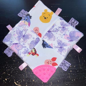 May include: A baby sensory toy featuring Winnie the Pooh characters and butterfly patterns. The square toy has a pink silicone corner, pink ribbons, and lace trim. The fabric features images of Pooh, Piglet, Eeyore, and Tigger.