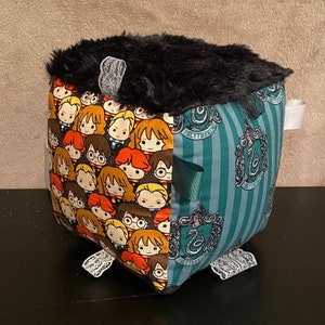May include: A black and green fabric cube with a black fur top. The cube is decorated with images of Harry Potter characters and the Slytherin house crest.