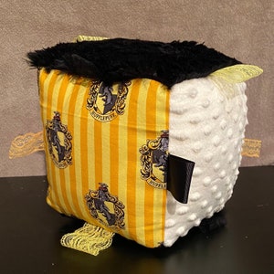 May include: A plush cube featuring a black faux fur top and sides with yellow and black striped fabric. The fabric is printed with the Hufflepuff crest. The cube has a white, textured side and yellow lace accents.