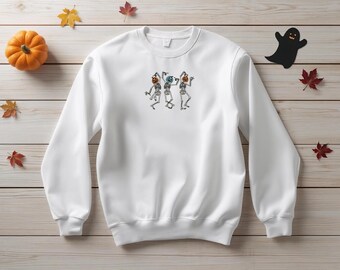 May include: White crewneck sweatshirt with an embroidered design of three dancing skeletons wearing pumpkin heads. The image includes a small orange pumpkin, a black ghost cutout, and autumn leaves on a wooden background. Halloween themed apparel.