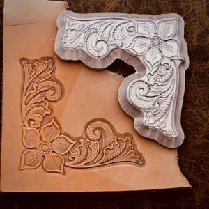 May include: A leathercraft corner design with a floral pattern is stamped onto a piece of tan leather. A metal stamp with the same floral design rests on the leather. The design includes flowers, leaves, and swirling patterns.