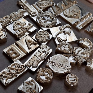 Puede incluir: Una colección de varios sellos metálicos con diferentes diseños y textos. Los sellos tienen varias formas, incluyendo rectangulares, circulares e irregulares. Algunos sellos presentan letras, mientras que otros tienen logotipos y elementos decorativos.
