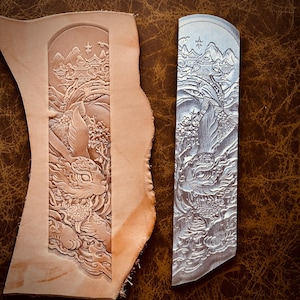 May include: A rectangular metal stamping tool featuring an intricate relief carving of a rabbit amidst clouds and traditional architecture, positioned next to a piece of light tan leather showing the same embossed design in high detail.