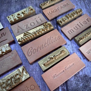 May include: A collection of leather stamps and brass letter stamps. The leather stamps are rectangular and embossed with names and words. The brass stamps are in various fonts and sizes. The background is a dark wood surface.
