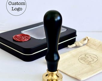 Può includere: Un timbro di ceralacca nero con base dorata e manico in legno nero. L'immagine mostra anche una scatola nera con un sigillo di cera rosso, un piccolo sacchetto e il testo "Custom Logo".