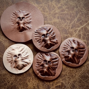 May include: A collection of brown leather coasters with embossed skull designs. The coasters vary in size and shade, with a detailed skull face in the center of each. The leather has a natural, textured appearance.