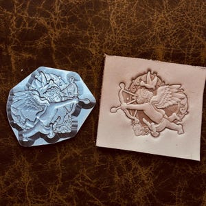 May include: A clear, hexagonal stamp with a detailed design of Cupid aiming a bow and arrow. Next to it, a piece of leather shows the embossed image. The design includes wings, a bow, and decorative elements.