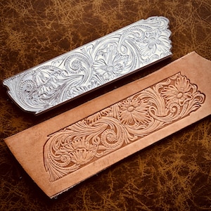 Puede incluir: Una herramienta de estampado de cuero de metal plateado con un diseño floral está junto a un trozo de cuero marrón estampado. La herramienta tiene un patrón detallado de flores y hojas. El cuero tiene un patrón floral en relieve similar.