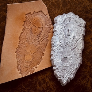 May include: A detailed leather carving and its corresponding stamp. The design features a woman's face above a snarling wolf. The leather is a light brown color, while the stamp is a light gray. The carving is on a piece of leather.