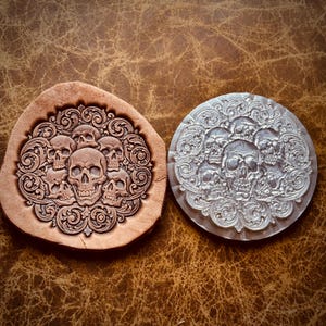 May include: A tan leather piece with a stamped design of five skulls surrounded by intricate scrollwork, positioned next to a clear circular acrylic stamp featuring the same skull and scrollwork pattern, both displaying identical detailed engravings.