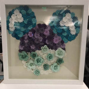 May include: A framed artwork featuring a Mickey Mouse silhouette made of paper flowers. The flowers are in shades of blue, purple, and green, creating a colorful and whimsical design.