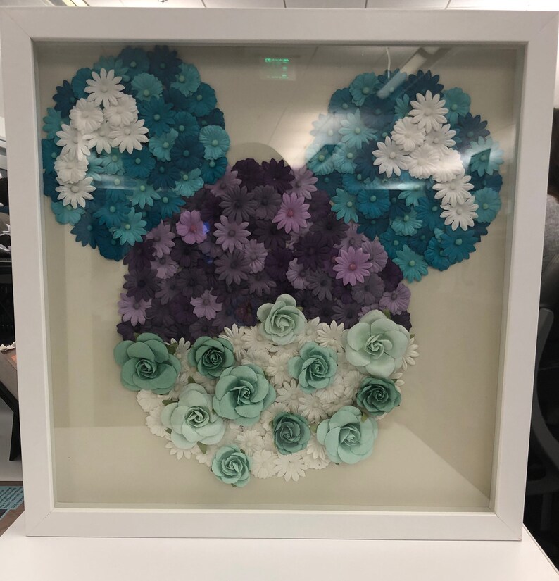 May include: A framed artwork featuring a Mickey Mouse silhouette made of paper flowers. The flowers are in shades of blue, purple, and green, creating a colorful and whimsical design.