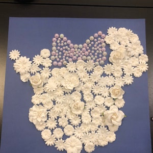 May include: A blue background with a white floral design in the shape of a mouse head. The mouse head is decorated with a purple bow made of pearls.