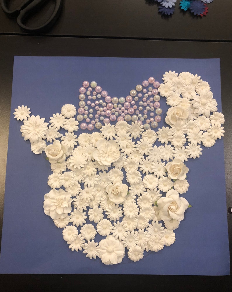 May include: A blue background with a white floral design in the shape of a mouse head. The mouse head is decorated with a purple bow made of pearls.
