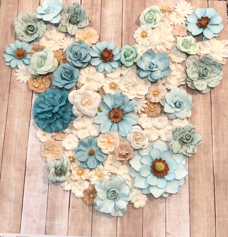 May include: A blue and white floral arrangement in the shape of a Mickey Mouse head. The flowers are made of paper and are arranged on a wooden background.