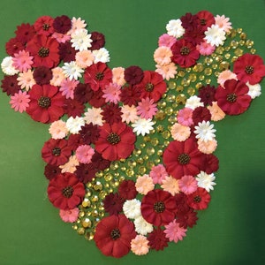 May include: A red and pink flower arrangement in the shape of Mickey Mouse. The flowers are surrounded by gold glitter.