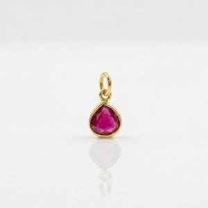 May include: A teardrop-shaped pendant featuring a faceted deep pink gemstone set in a polished gold-toned metal frame. The pendant includes a small circular jump ring at the top for attachment, showcasing a clear reflection on the surface below.