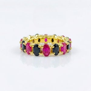 May include: A gold eternity band featuring an alternating pattern of oval-cut vibrant pink rubies and deep black sapphires. The gemstones are set in a continuous row within a polished gold metal setting, creating a circular ring design.