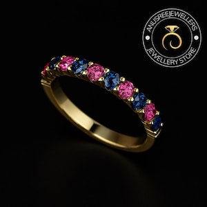 May include: A gold ring featuring a row of alternating pink and blue gemstones. The ring is set against a black backdrop, accentuating the colours of the stones. The ring is from Anusree Jewellers Jewellery Store.