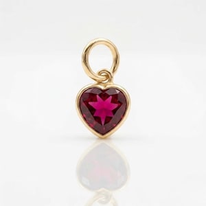 May include: A gold heart-shaped pendant featuring a deep red faceted gemstone set in a polished gold frame. The pendant includes a circular gold jump ring at the top for attachment, showcasing a vibrant crimson hue and a clear, reflective surface.