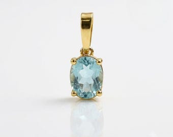 Puede incluir: Un colgante de oro con una piedra preciosa de color azul claro de corte ovalado sujeta por cuatro garras. El colgante incluye una anilla de oro pulido en la parte superior para engancharlo a una cadena, mostrando una piedra clara y facetada.