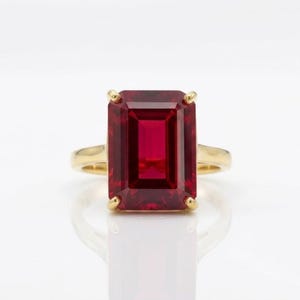 May include: A gold ring featuring a large, rectangular emerald-cut deep red gemstone set in a four-prong setting. The band is smooth and polished gold, providing a clean, classic look that highlights the vibrant color and clarity of the stone.
