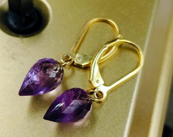 May include: A pair of gold-colored earrings with teardrop-shaped, faceted amethyst gemstones. The earrings have a lever-back closure and are displayed on a gold-toned surface. The amethyst stones have a deep purple hue.