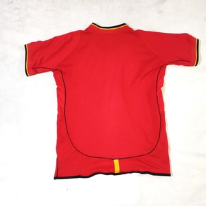 K&ouml;nnte beinhalten: Rotes Kurzarm-Trikot mit schwarzem und gelbem Besatz. Das Trikot hat einen schwarzen Kragen und einen schwarzen und gelben Streifen in der Mitte des R&uuml;ckens.