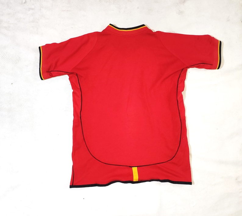 K&ouml;nnte beinhalten: Rotes Kurzarm-Trikot mit schwarzem und gelbem Besatz. Das Trikot hat einen schwarzen Kragen und einen schwarzen und gelben Streifen in der Mitte des R&uuml;ckens.