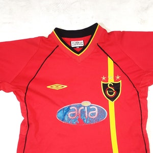 K&ouml;nnte beinhalten: Rotes Fu&szlig;balltrikot mit schwarzem und gelbem Besatz. Das Trikot hat einen schwarzen Kragen, einen gelben Streifen auf der rechten Seite und ein Logo auf der linken Brust. Das Logo ist eine Raute mit gelbem Rand. Das Trikot hat auch ein Logo auf der rechten Brust, das ein Schild mit schwarzem Rand und einem gelben Stern auf jeder Seite ist. Das Schild hat einen schwarzen Buchstaben "S" darin. Das Trikot hat ein blaues und wei&szlig;es ovales Logo mit dem Wort "aria" in wei&szlig;.