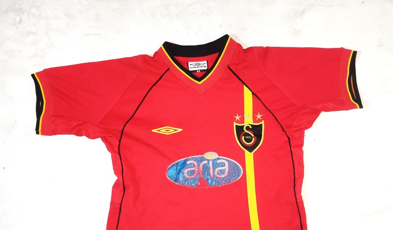 K&ouml;nnte beinhalten: Rotes Fu&szlig;balltrikot mit schwarzem und gelbem Besatz. Das Trikot hat einen schwarzen Kragen, einen gelben Streifen auf der rechten Seite und ein Logo auf der linken Brust. Das Logo ist eine Raute mit gelbem Rand. Das Trikot hat auch ein Logo auf der rechten Brust, das ein Schild mit schwarzem Rand und einem gelben Stern auf jeder Seite ist. Das Schild hat einen schwarzen Buchstaben "S" darin. Das Trikot hat ein blaues und wei&szlig;es ovales Logo mit dem Wort "aria" in wei&szlig;.