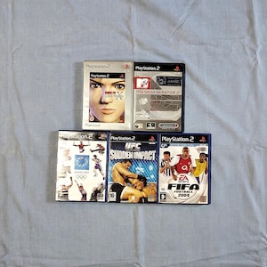 May include: A collection of six PlayStation 2 video game cases. The cases are all silver and blue with various game titles including "Music Generator 2", "UFC Sudden Impact", and "FIFA Football 2004".