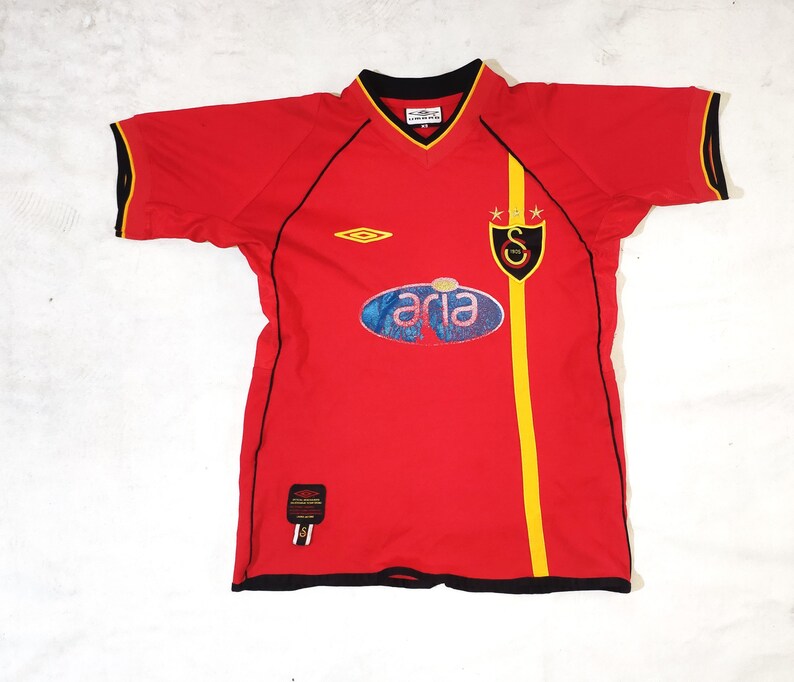 K&ouml;nnte beinhalten: Rotes Fu&szlig;balltrikot mit schwarzem und gelbem Besatz. Das Trikot hat ein schwarz-wei&szlig;es Logo auf der linken Brust, ein schwarz-goldenes Logo auf der rechten Brust und ein blau-wei&szlig;es Logo auf der Vorderseite. Der Text "aria" befindet sich auf dem blau-wei&szlig;en Logo.