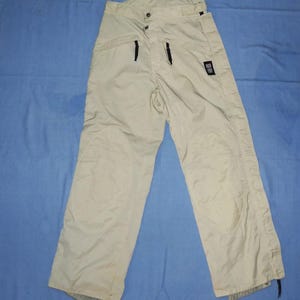Op de afbeelding: Beige sneeuwbroek met zwarte ritsen en een kleine zwarte logopatch. De broek heeft verstevigde kniepanelen en een knoop- en ritssluiting in de taille. Ontworpen voor wintersport en buitenactiviteiten.