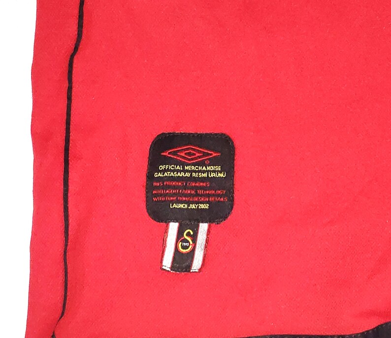 K&ouml;nnte beinhalten: Ein schwarz-wei&szlig;es Etikett mit dem Text "OFFICIAL MERCHANDISE GALATASARAY RESMİ &Uuml;R&Uuml;N&Uuml;" und "LAUNCH JULY 2002". Das Etikett ist an einem roten Shirt befestigt.