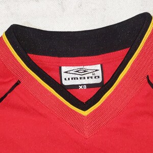 K&ouml;nnte beinhalten: Rotes Fu&szlig;balltrikot mit schwarzem und gelbem Besatz am Hals. Das Trikot hat ein wei&szlig;es Logo mit dem Text "UMBRO" und der Gr&ouml;&szlig;e "XS".