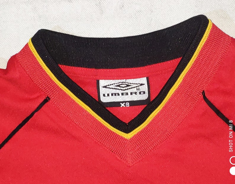 K&ouml;nnte beinhalten: Rotes Fu&szlig;balltrikot mit schwarzem und gelbem Besatz am Hals. Das Trikot hat ein wei&szlig;es Logo mit dem Text "UMBRO" und der Gr&ouml;&szlig;e "XS".