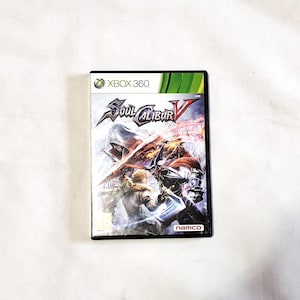以下が含まれることがあります： Xbox 360用ゲームケース、ソウルキャリバーV。赤と白の背景に、戦闘中のキャラクターが描かれている。ゲームケースは黒で、白い縁取りと白い文字で「Xbox 360」と「ソウルキャリバーV」と書かれている。