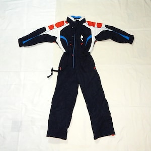 Op de afbeelding: Marineblauwe en witte skipak met rode accenten. Het eendelige pak heeft een hoge kraag, een ritssluiting aan de voorkant en een zwarte riem. De mouwen hebben blauwe en witte details. Het pak is ontworpen voor wintersport en buitenactiviteiten.