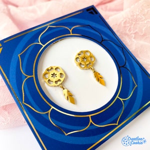 Puede incluir: Pendientes de color dorado con un diseño circular delicado y un colgante de pluma. Los pendientes se muestran sobre un fondo azul con un borde dorado.