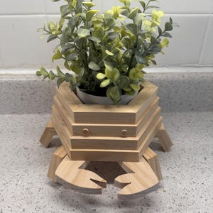 May include: A wooden crab-shaped planter with a white pot holding green and yellow artificial plants. The crab planter is made of light-colored wood and has a layered design. The crab's claws and legs are also made of wood.