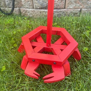May include: A bright red wooden crab-shaped garden stake. The crab has a hexagonal body, two large claw-like appendages, and a vertical pole extending upwards. The stake is painted a vibrant red and sits on green grass.